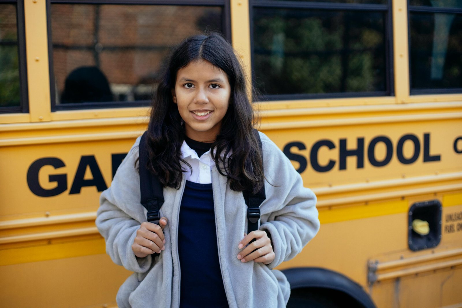 Happy Hispanic girl with backpack smiling in front of a yellow school bus, ready for school.